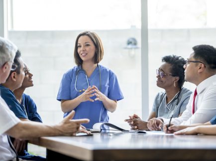 A multi-ethnic group of medical staff are indoors in a hospital. They are wearing medical clothing. A Caucasian female doctor is giving a presentation to the others.